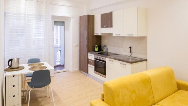 A compact kitchen with a stove, oven, and sink, a dining area with a table and chairs, and a yellow sofa. - RoomApartment Rome: 1 Bedroom Apartment with Balcony  (Roma)