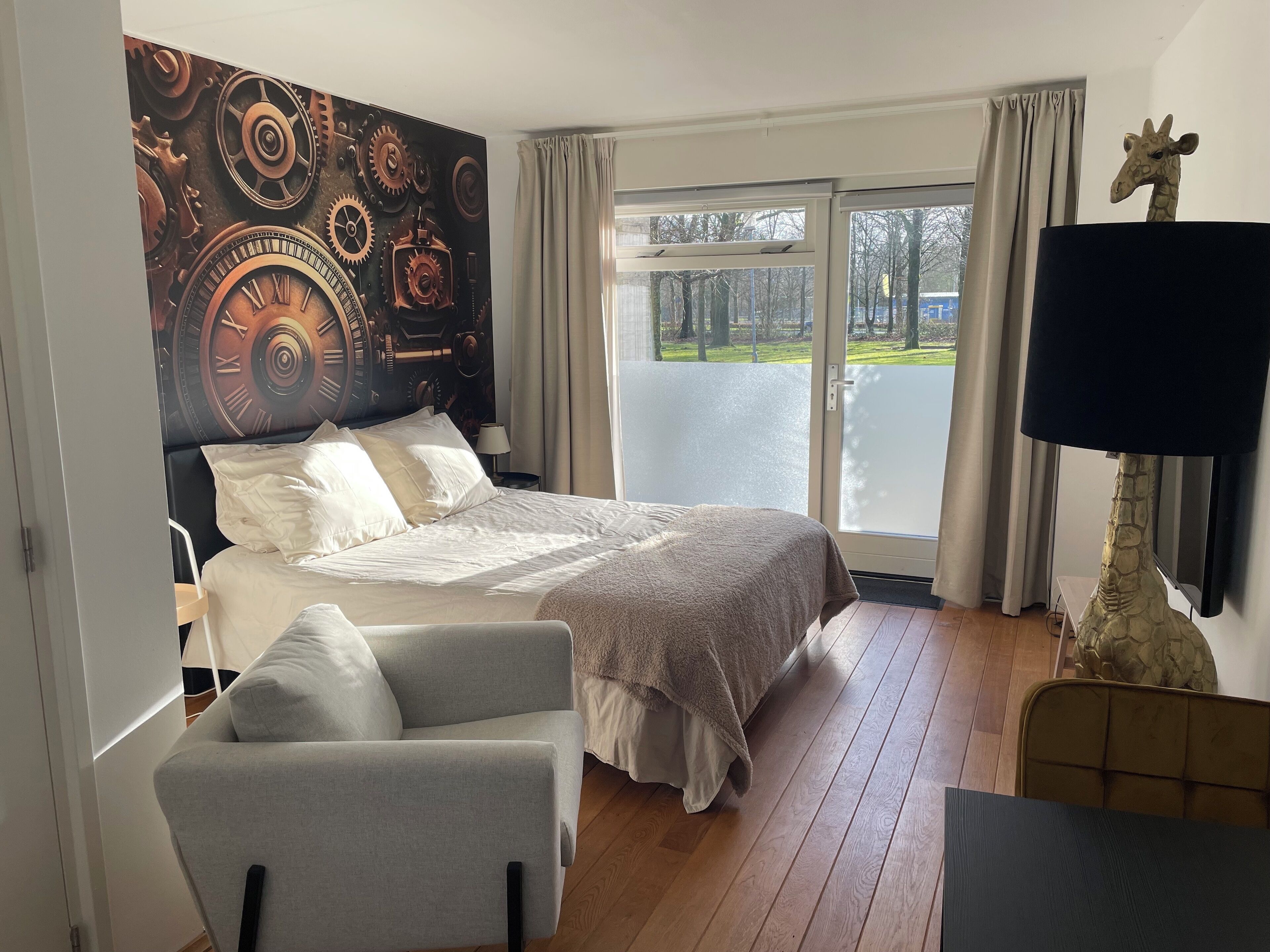 A bedroom with a large bed, a chair, a giraffe decoration, and a view of the outdoors through a sliding door.