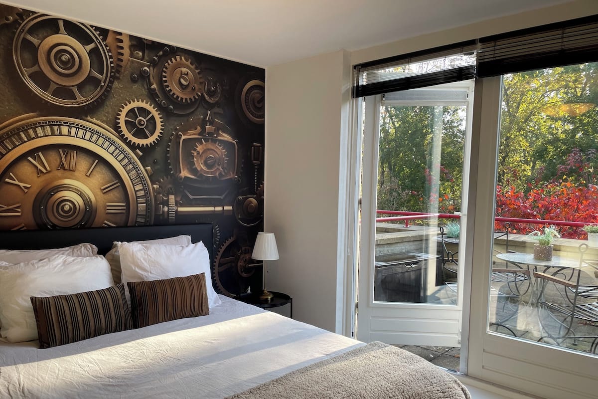A hotel room with a large bed, a clock-themed wall mural, a window with a view of a garden and outdoor seating.