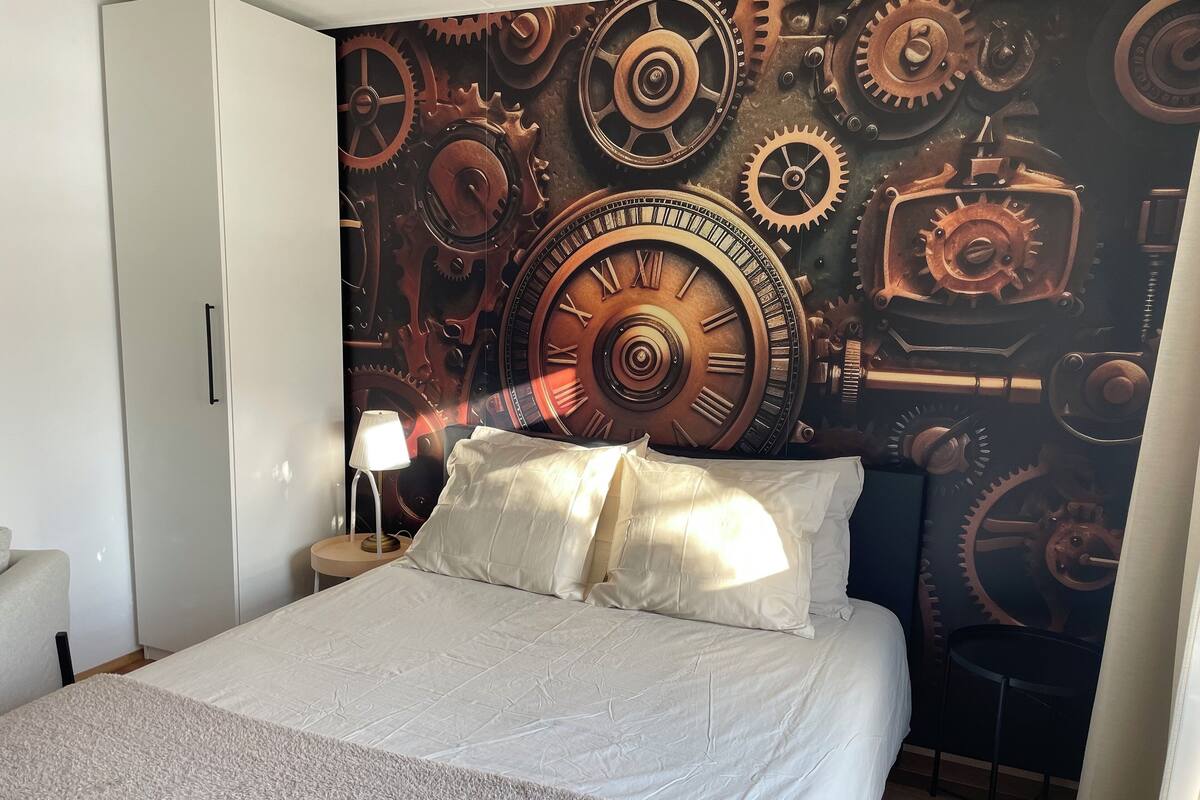 A bedroom with a large bed, a nightstand with a lamp, and a wall with a mechanical gear design.