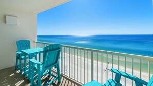 Un balcon avec des chaises turquoise et une table donnant sur une plage aux eaux bleu cristallin et au rivage sablonneux.