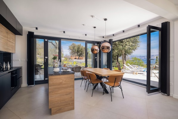 Une cuisine moderne dotée d’un grand îlot central, d’un coin repas avec des chaises et de grandes fenêtres offrant une vue sur l’extérieur.