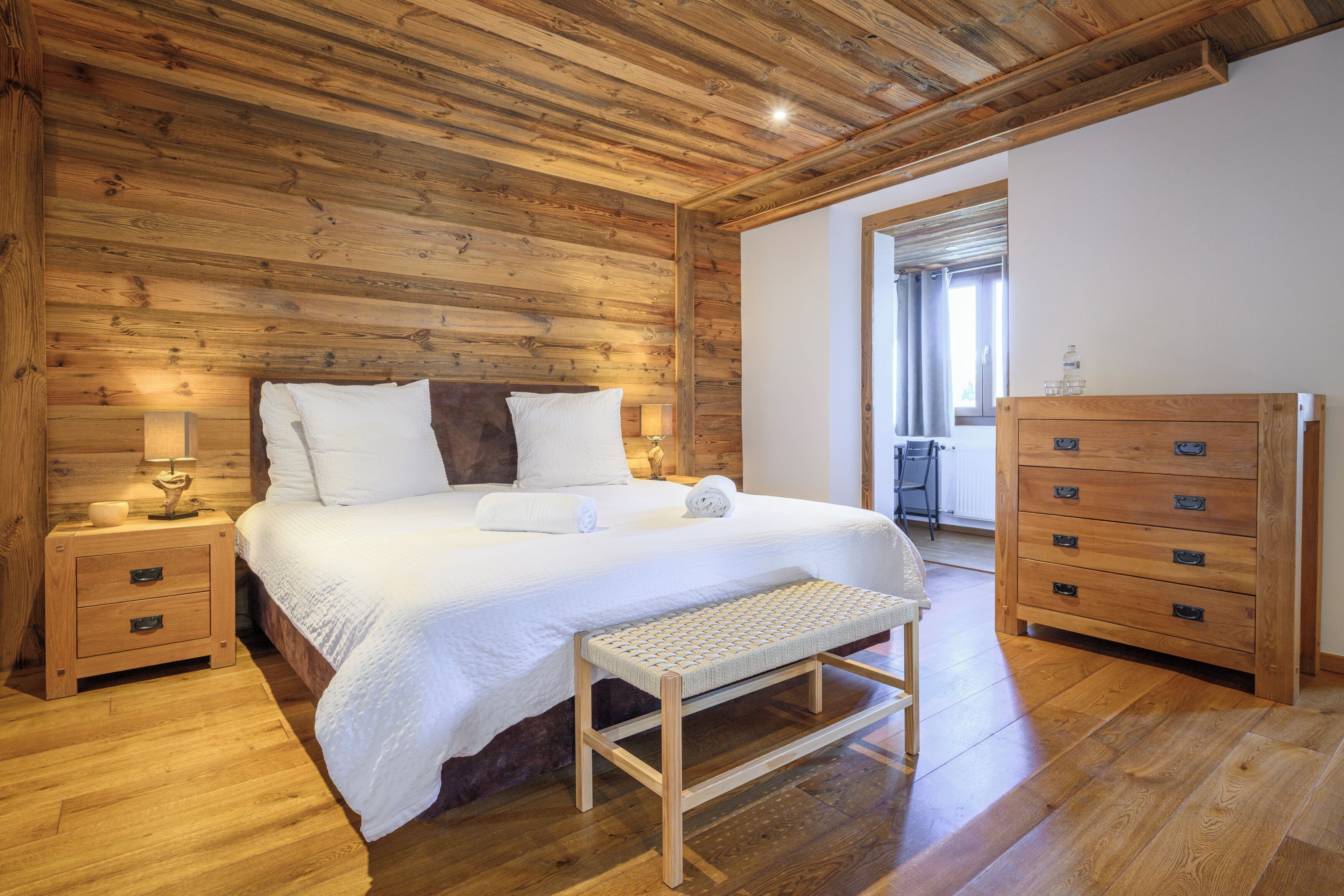 Une chambre aux murs lambrissés, avec un lit, des tables de chevet, un banc et une commode en bois.