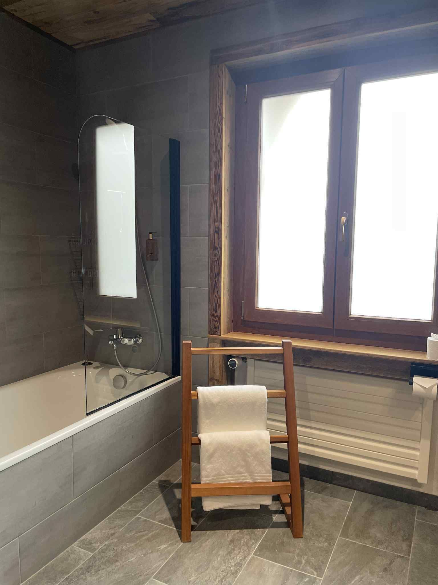 Un baño con bañera, mampara de vidrio, una silla de madera con una toalla y una ventana con persianas.