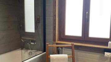 A bathroom with a bathtub, a glass shower enclosure, a wooden chair with a towel, and a window with blinds.