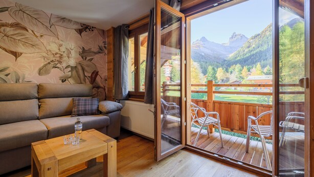 A cozy room with a sofa, a wooden table, and a sliding glass door leading to an outdoor deck with chairs. - Hôtel des Muverans (Leytron)