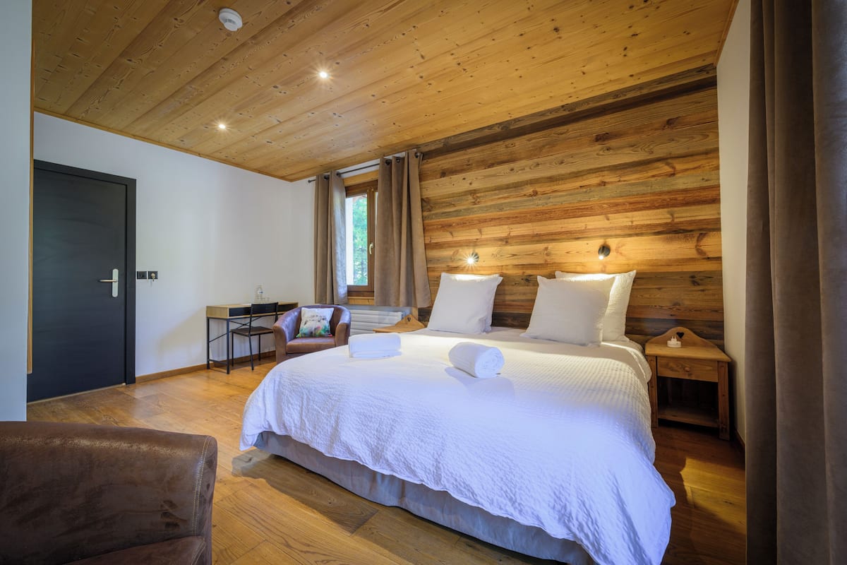 A bedroom with a wooden ceiling, a bed with white linens, a desk with a chair, and a window with curtains.