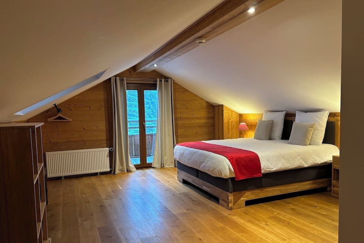 A bedroom with a sloped ceiling, wooden flooring, a bed with white bedding and a red throw, a radiator, and a sliding glass door leading to a balcony.