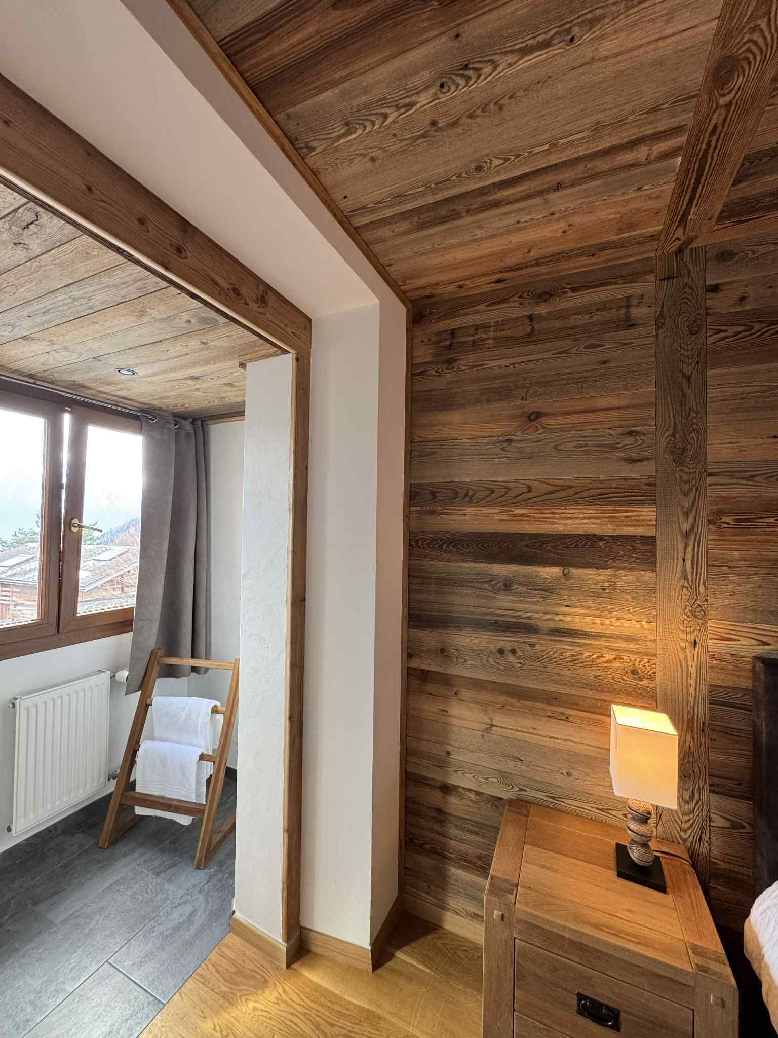 A room with wooden walls and ceiling, a wooden nightstand with a lamp, a wooden ladder, and a window with curtains.