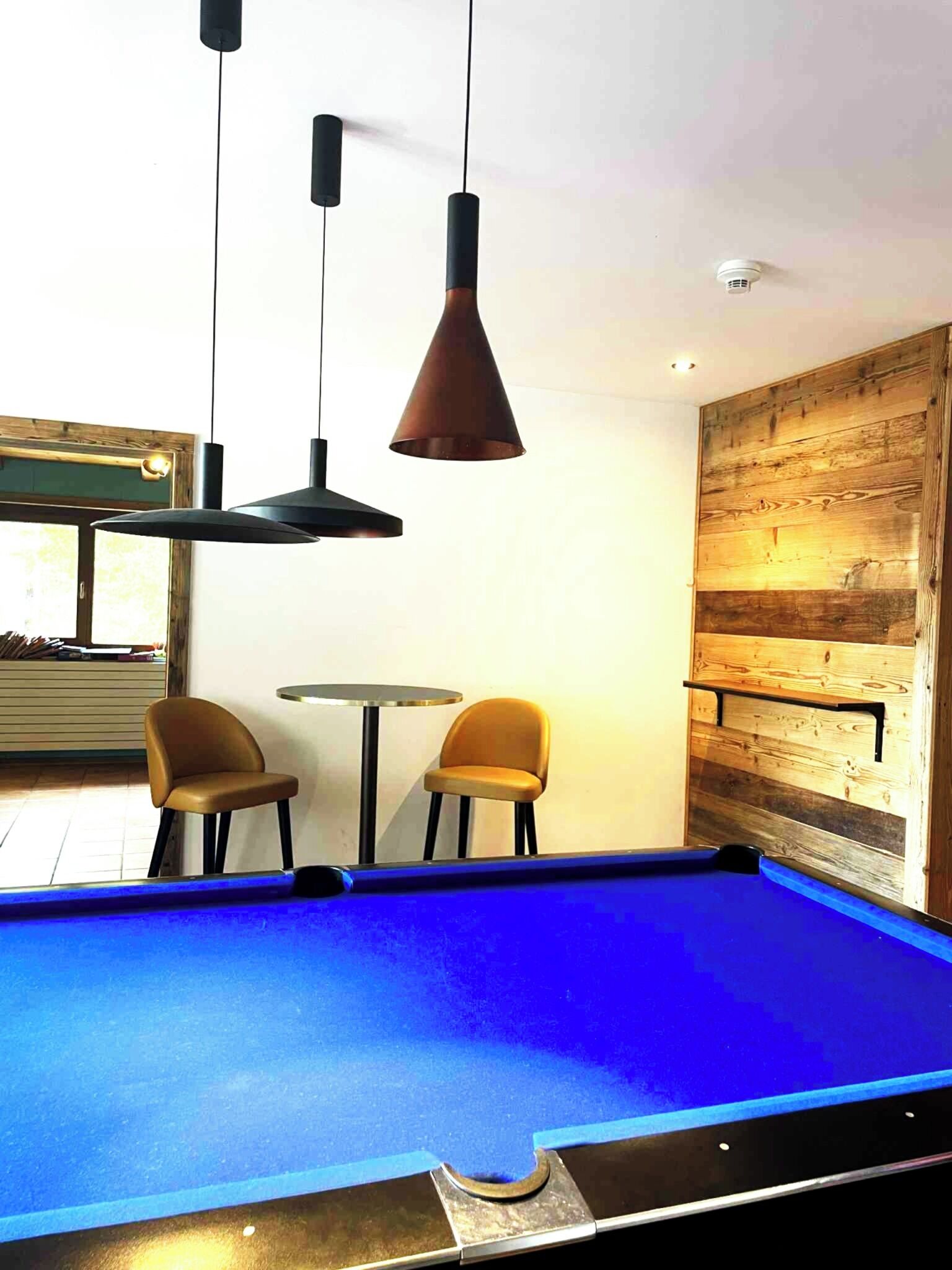 Een pooltafel met twee stoelen, een kleine ronde tafel en hanglampen in een kamer met houten lambrisering en een raam.