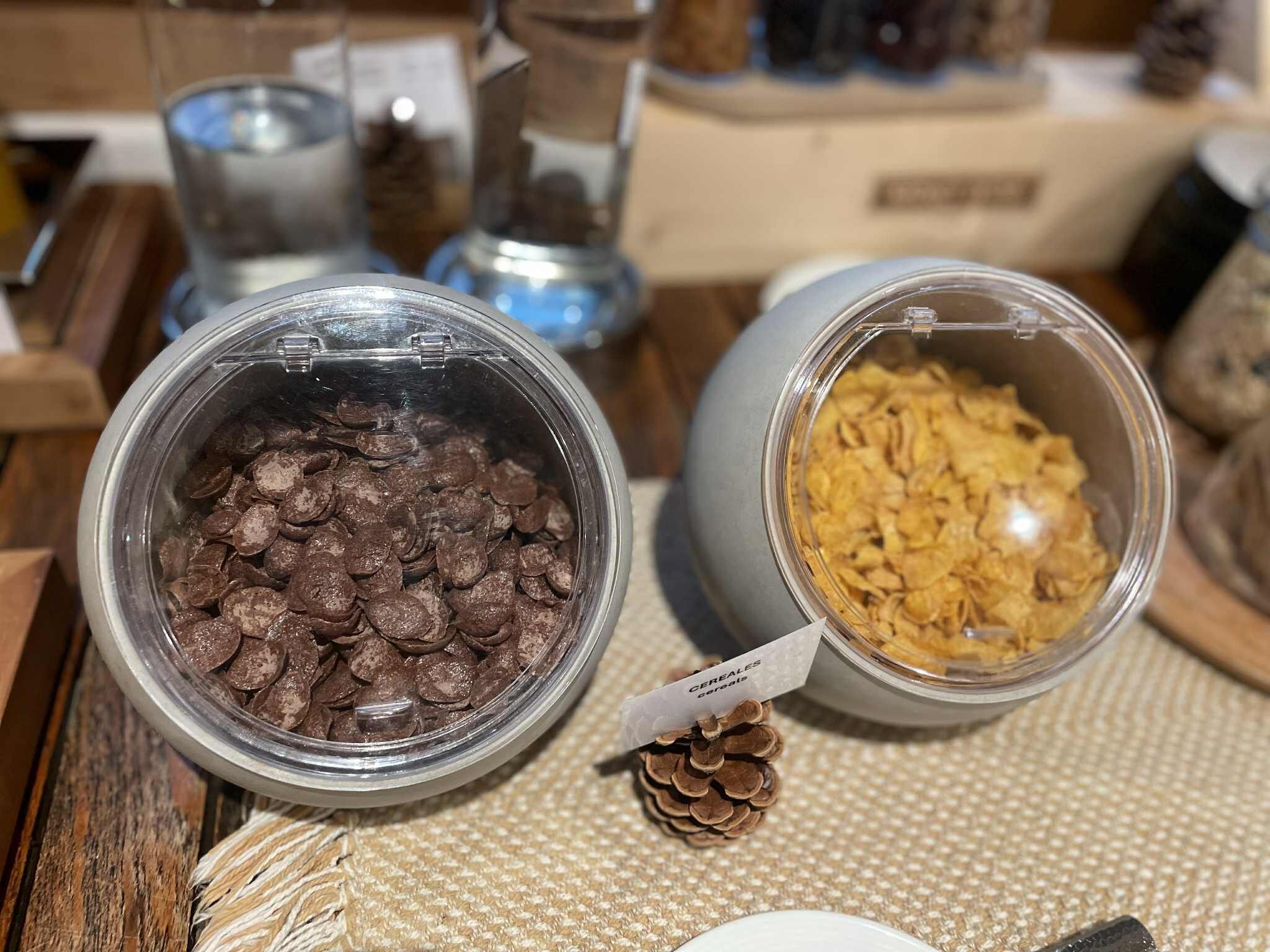 Two glass jars on a woven mat, one filled with chocolate chips and the other with cereal flakes, with a pine cone and a label between them.