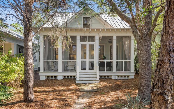 A white house with a covered porch, surrounded by trees and a wooden deck.