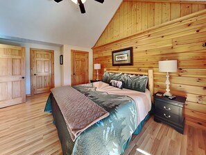 Une chambre avec un plafond en bois, un lit recouvert d’une couvre-lit à motifs, deux tables de chevet avec des lampes et une armoire en bois.