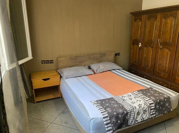 A bedroom with a bed, a wooden nightstand, a wardrobe, and a tiled floor.