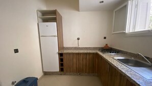 Een kleine keuken met houten kastjes, een witte koelkast, een spoelbak en een raam met jaloezieën.