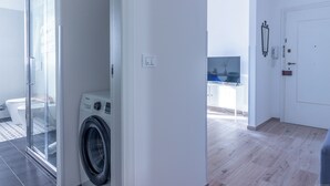 Eine moderne Wohnung mit Duschkabine aus Glas, einer Waschmaschine, einer weißen Tür und einem sichtbaren Küchenbereich.