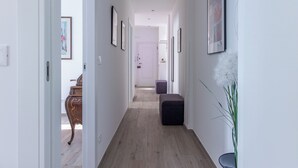 Ein schmaler Flur mit Holzboden, weißen Wänden und gerahmten Kunstwerken. Auf der rechten Seite befindet sich eine dunkle Bank und ein Blumentopf.