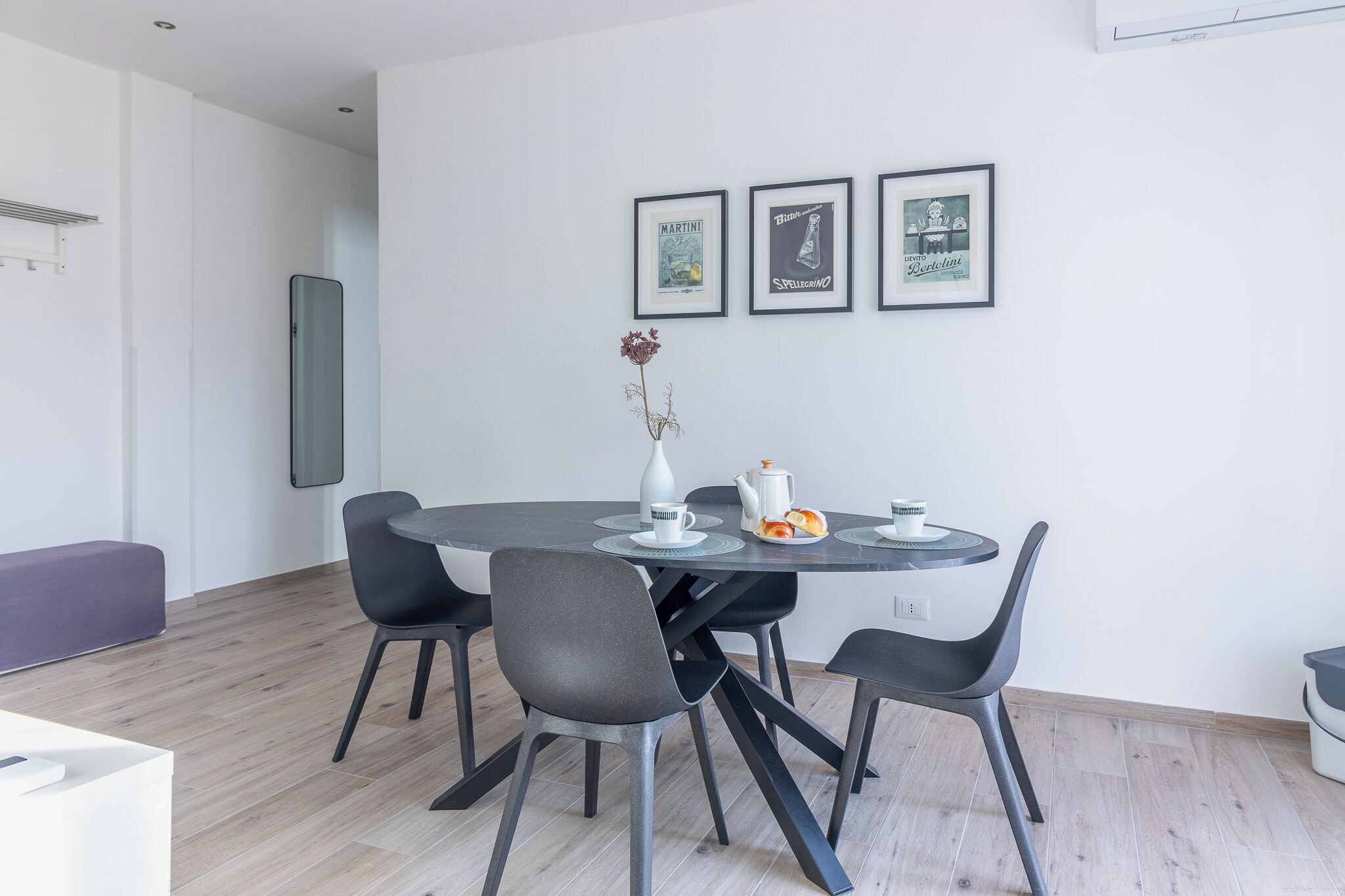 Un comedor moderno con una mesa redonda, cuatro sillas y un florero con flores. Hay tres cuadros enmarcados en la pared sobre la mesa.