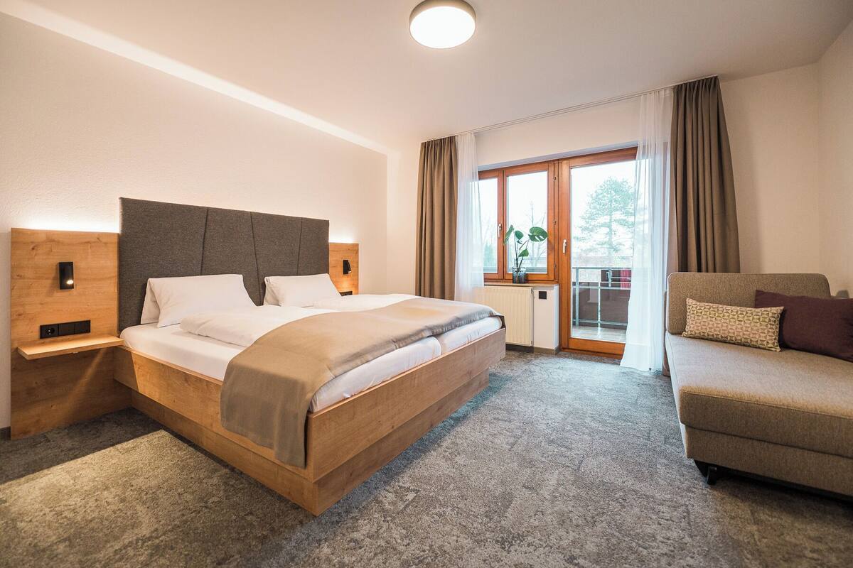 A hotel room with a wooden bed, a sofa, a window with curtains, and a view of the outdoors.