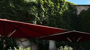 An outdoor dining area with tables and chairs under red umbrellas, surrounded by greenery and a stone wall. - Hotel Goldener Engel (Ihringen)