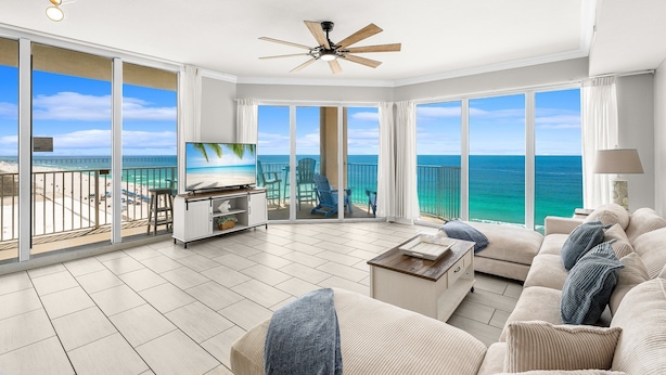 A spacious living room with a large TV, a ceiling fan, and a view of the ocean through large windows.