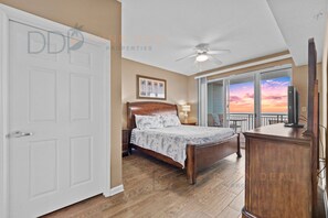 A bedroom with a bed, a nightstand, a dresser, a ceiling fan, and a view of the sunset through the window. - Legacy Tower II 509 | Deluxe Unit with Stunning Coastal Views! (Gulfport)