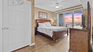 Een slaapkamer met een bed, een nachtkastje, een dressoir, een plafondventilator en uitzicht op de ondergaande zon door het raam.