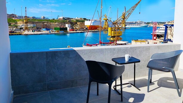 Un balcón con dos sillas y una mesa, con vistas a un puerto con grúas y un crucero.