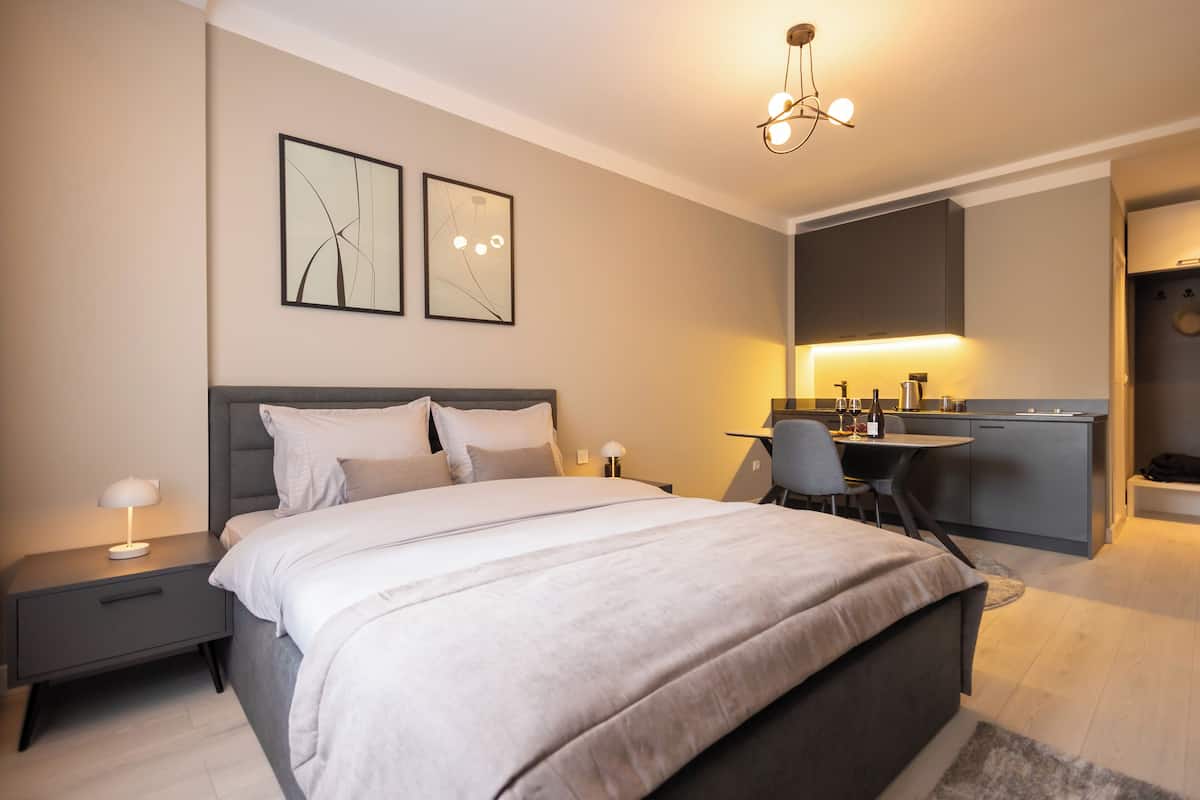 A modern bedroom with a large bed, bedside tables, a desk, and a kitchenette.