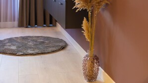 Una habitación de hotel moderna con un televisor de pantalla plana montado en la pared, un aparador de color oscuro, un tapete redondo en el suelo y un jarrón con flores secas sobre una mesita de noche.