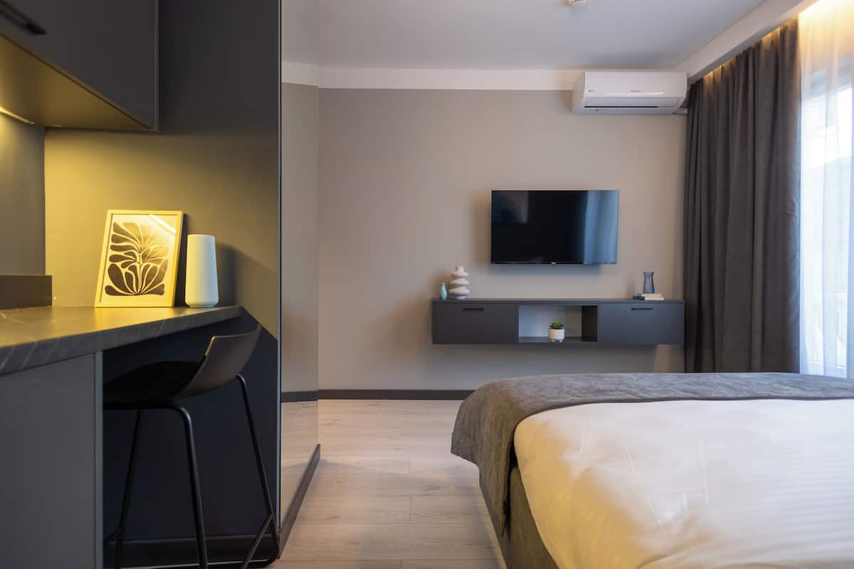A modern hotel room with a bed, a desk with a chair, a flat-screen TV, and a small shelf with decorative items.