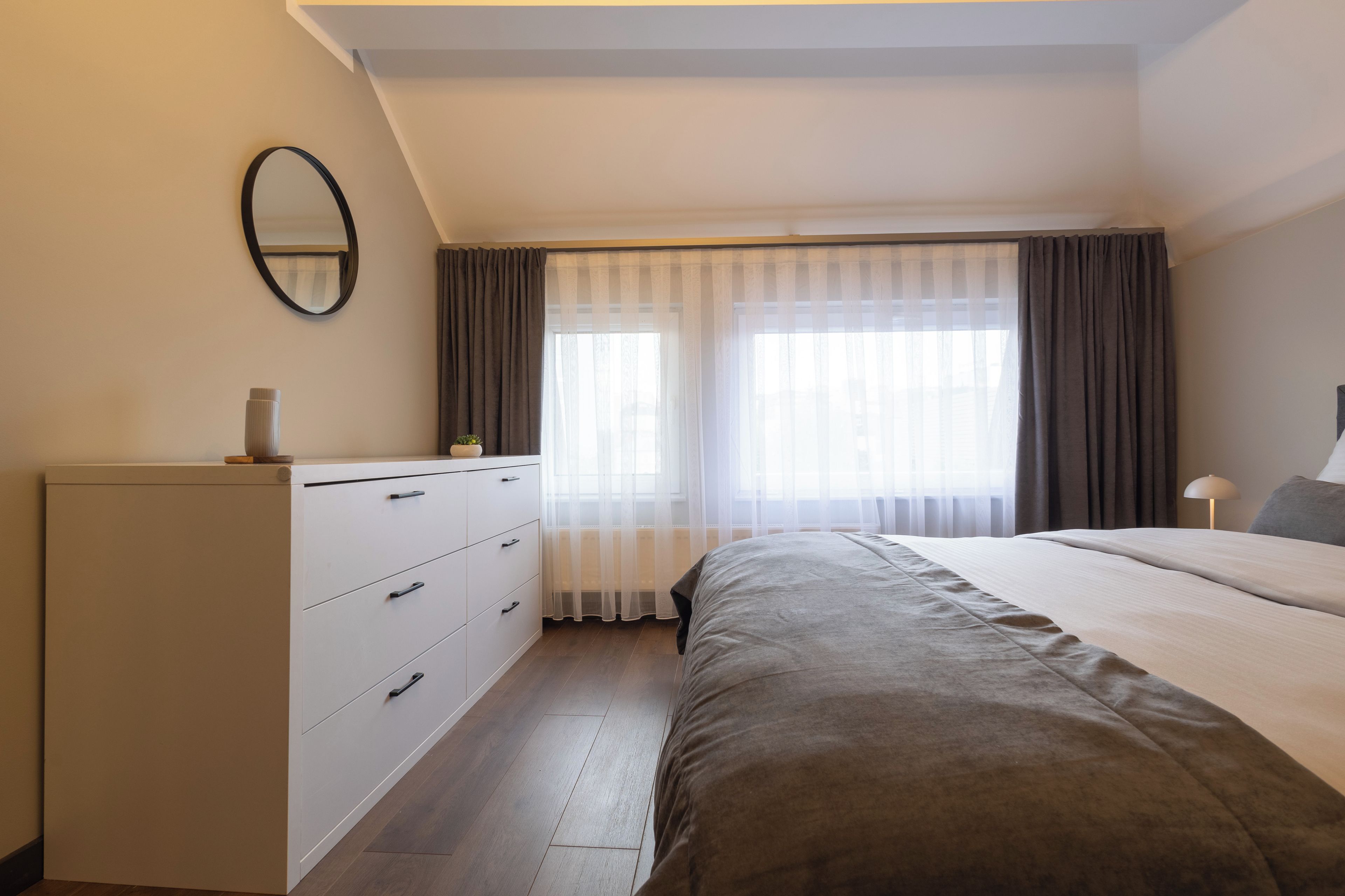 Een slaapkamer met een bed, een dressoir, een spiegel en een raam met gordijnen.