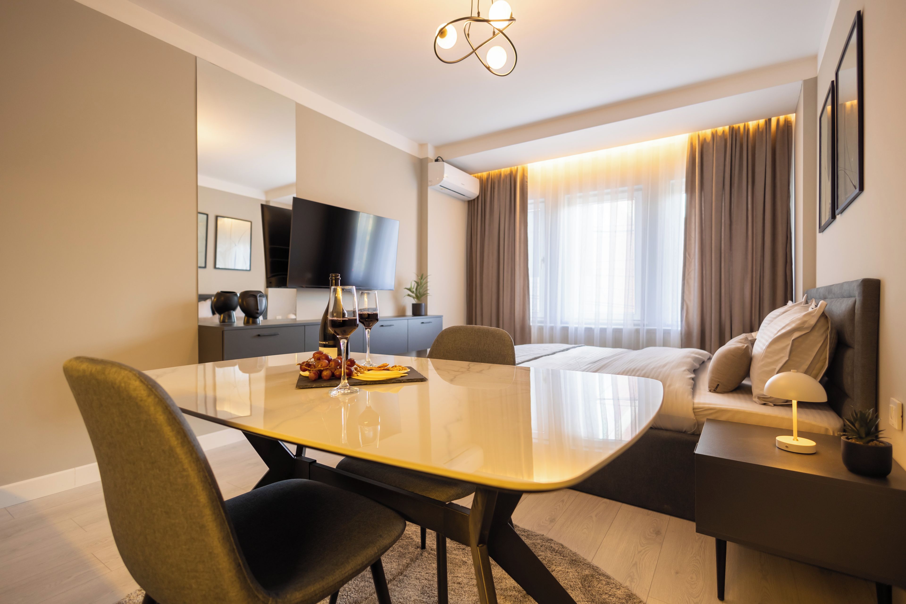 Ein modernes Hotelzimmer mit Esstisch, Stühlen, einer Sitzecke, einem Fernseher und einem Fenster mit Vorhängen.