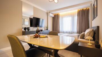 Ein modernes Hotelzimmer mit Esstisch, Stühlen, einer Sitzecke, einem Fernseher und einem Fenster mit Vorhängen.