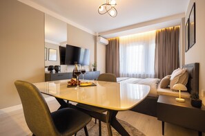 Ein modernes Hotelzimmer mit Esstisch, Stühlen, einer Sitzecke, einem Fernseher und einem Fenster mit Vorhängen.