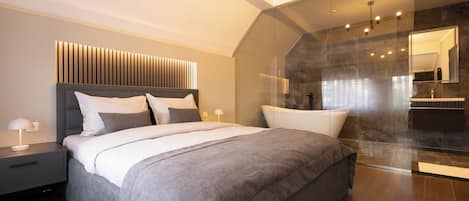 Quarto moderno com uma cama grande, mesa de cabeceira com abajur e área de chuveiro envidraçada.