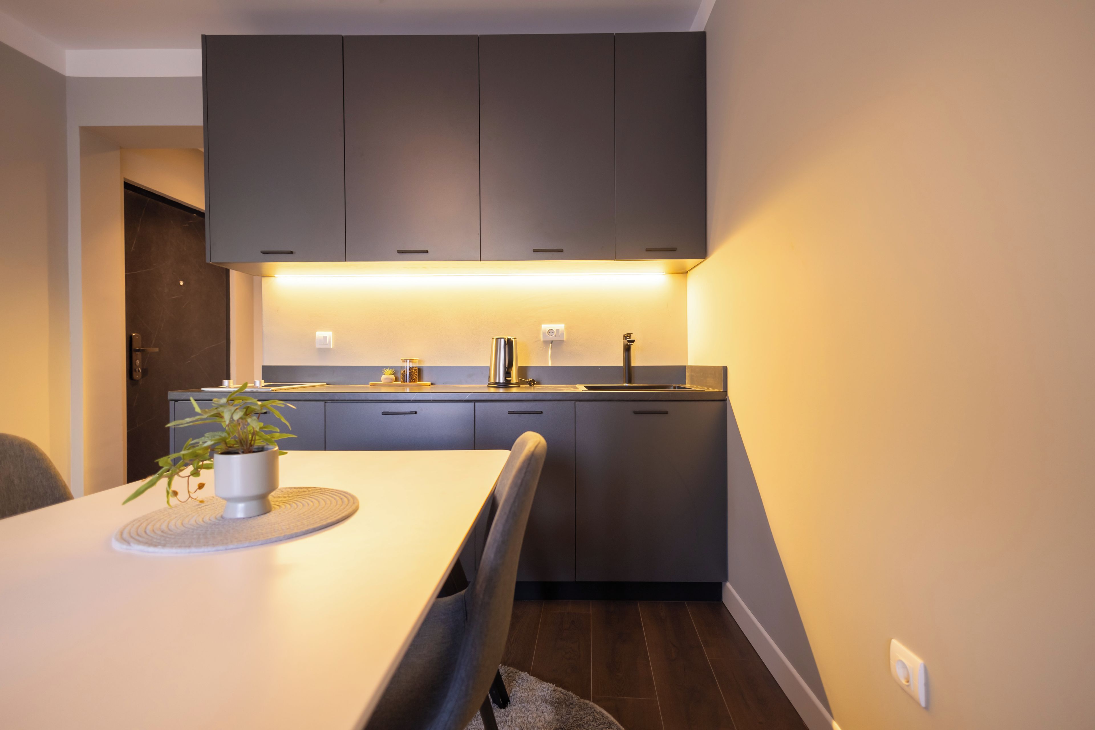 Een moderne keuken met donkere kastjes, een wit aanrechtblad en een eethoek met een tafel en stoelen.