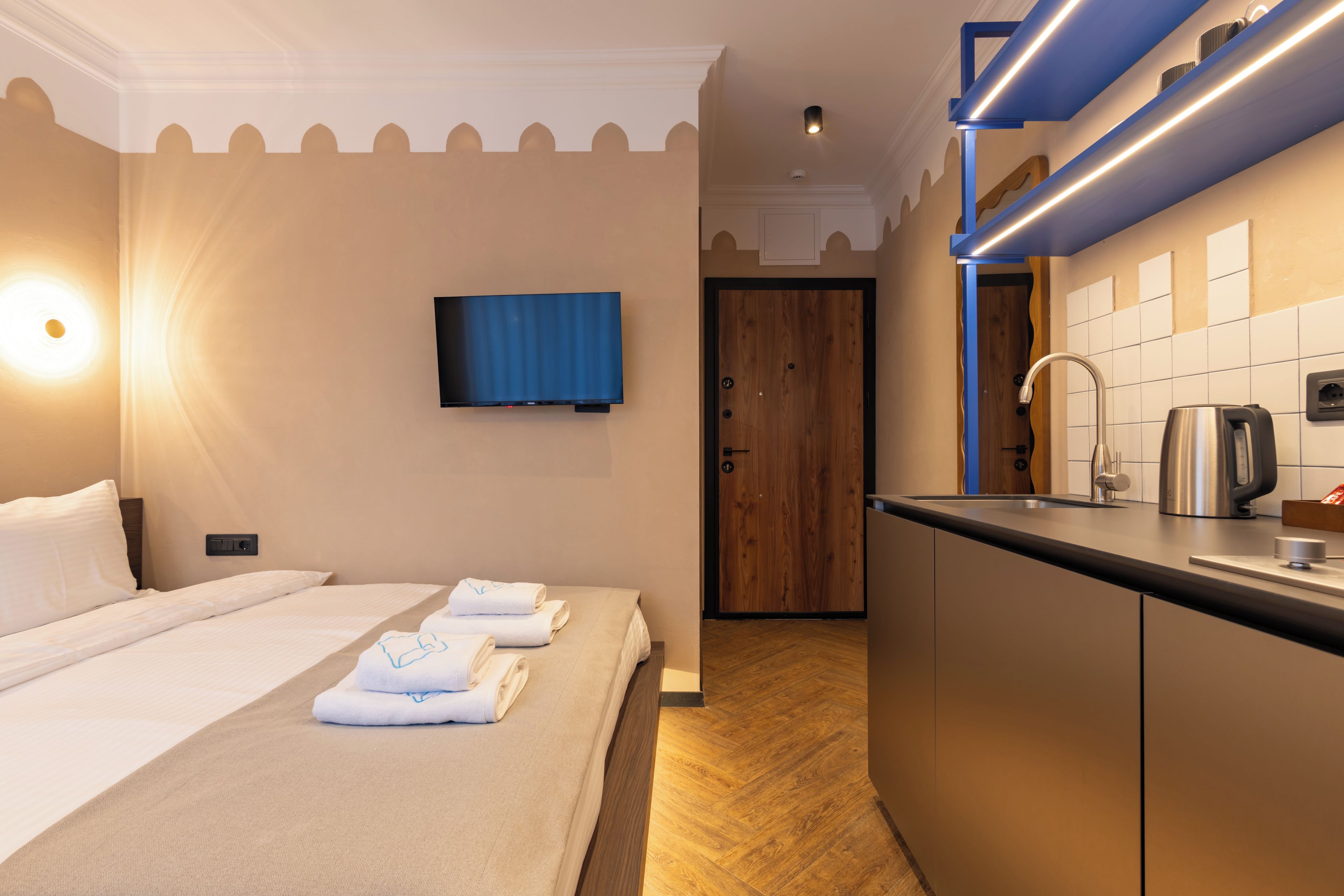 A hotel room with a bed, a TV mounted on the wall, a wooden door, and a kitchenette with a kettle.