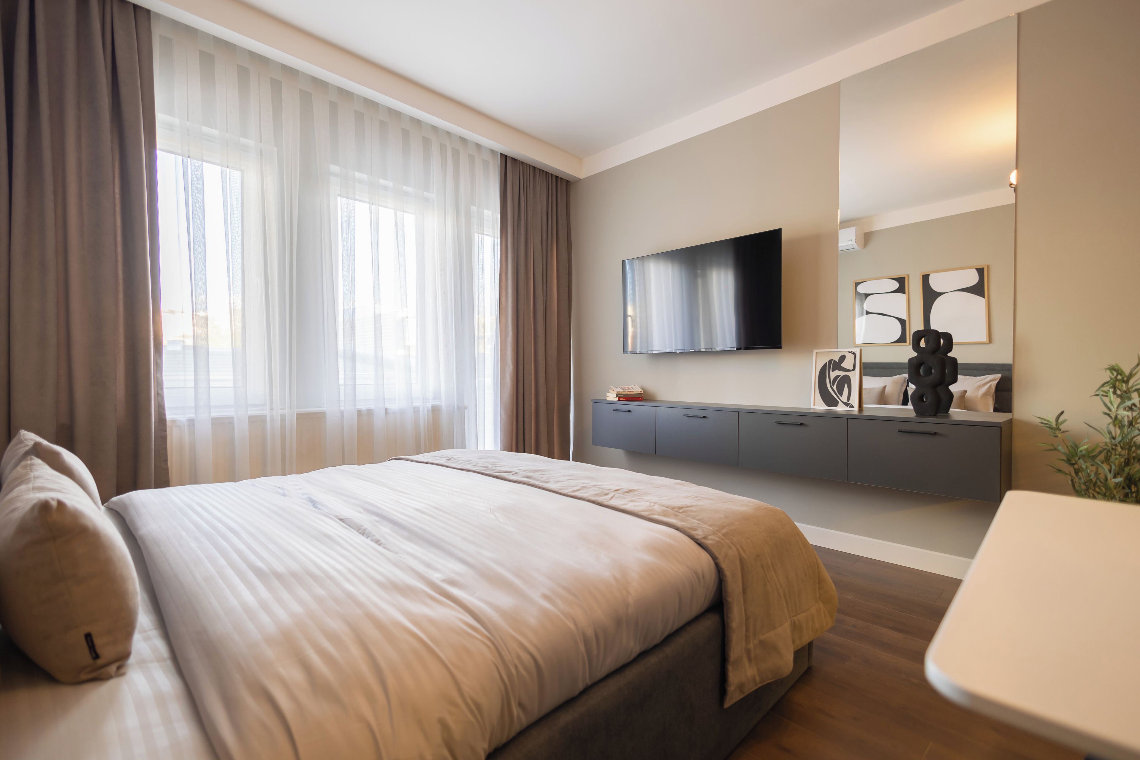 Una habitación de hotel moderna con una cama grande, un televisor de pantalla plana montado en la pared, una mesita de noche y una pequeña zona de escritorio.