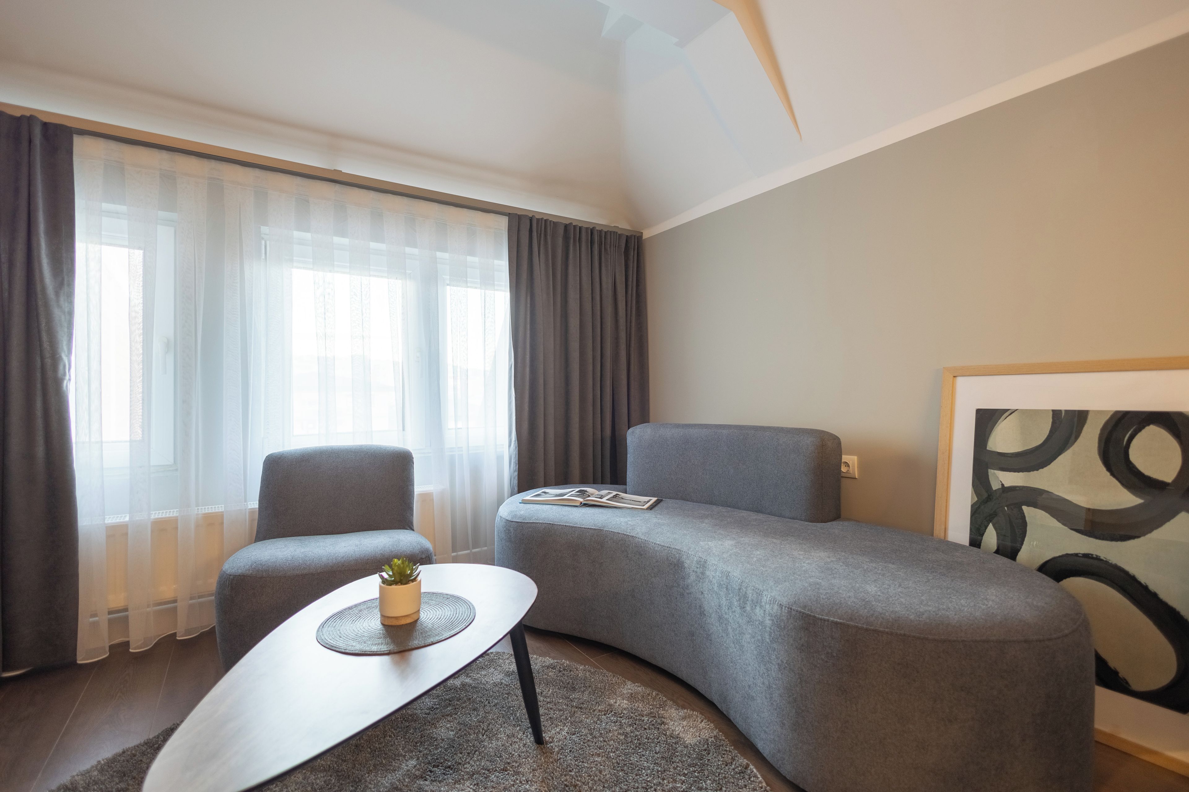 Een moderne woonkamer met een grijze bank, een klein bijzettafeltje en een groot raam met dunne gordijnen.