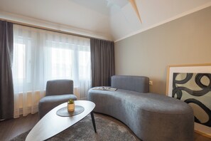 Una sala moderna con un sofá gris, una mesita de centro pequeña y un ventanal con cortinas traslúcidas.