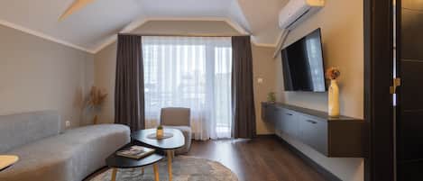 A modern living room with a sofa, a coffee table, a TV mounted on the wall, and a large window with curtains.