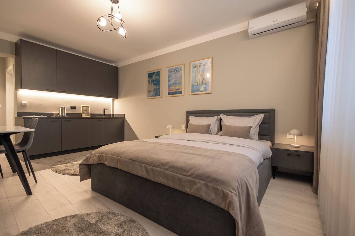 A modern bedroom with a large bed, bedside tables, and a small desk. There are framed pictures on the wall and an air conditioning unit above the bed.