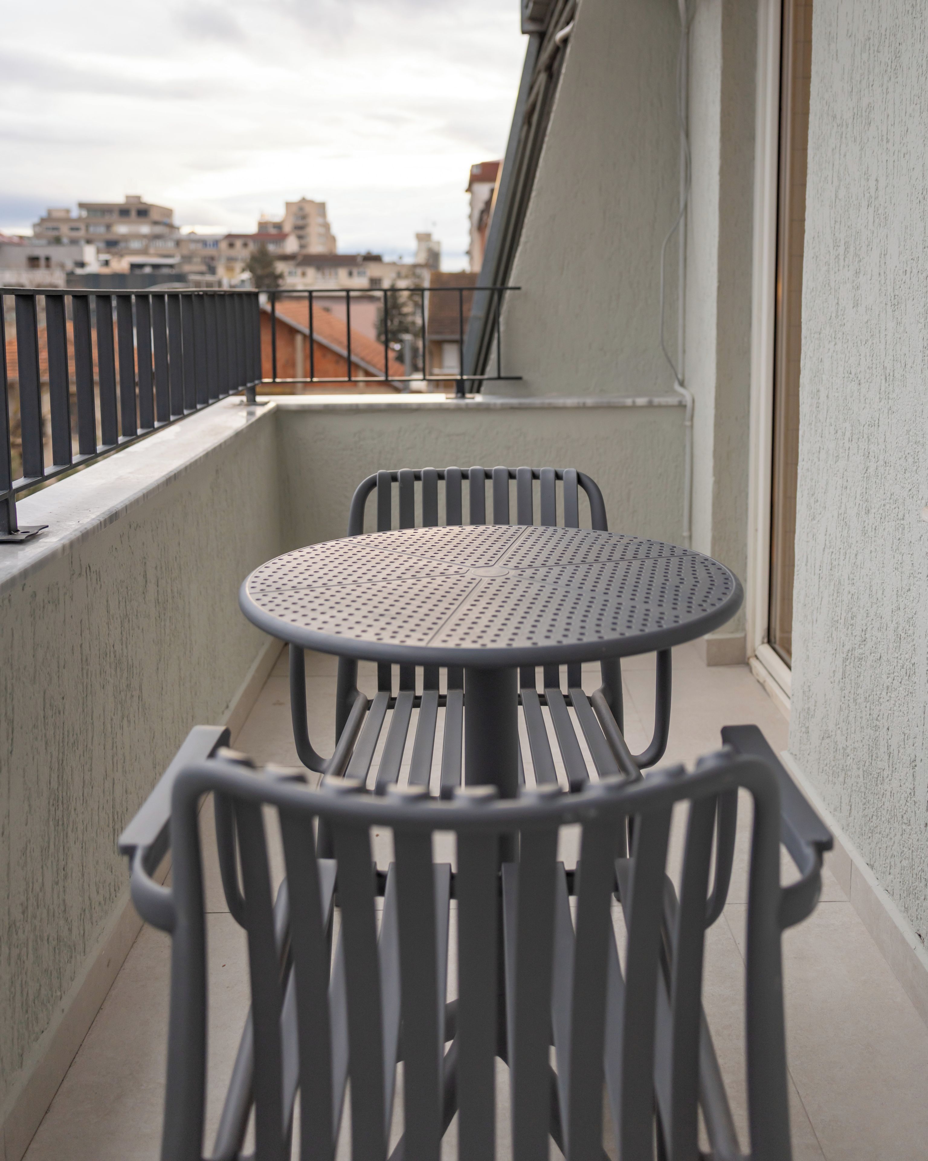 Een balkon met een ronde tafel en twee stoelen, een metalen hekwerk en een uitzicht op gebouwen op de achtergrond.
