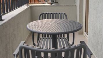 Een balkon met een ronde tafel en twee stoelen, een metalen hekwerk en een uitzicht op gebouwen op de achtergrond.