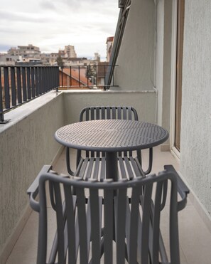 Un balcón con una mesa redonda y dos sillas, barandilla metálica y vistas a edificios al fondo.