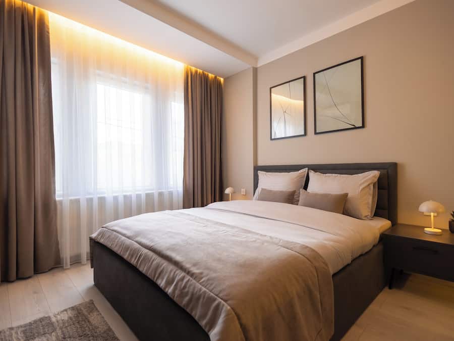 Um quarto moderno com uma cama grande, uma mesa de cabeceira com abajur e uma janela com cortinas.