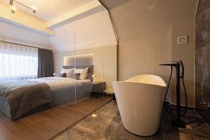 A modern hotel room with a large bed, a bedside table, a nightstand, a large white bathtub, and a floor lamp. - Prishtina City Apartments (Pristina)