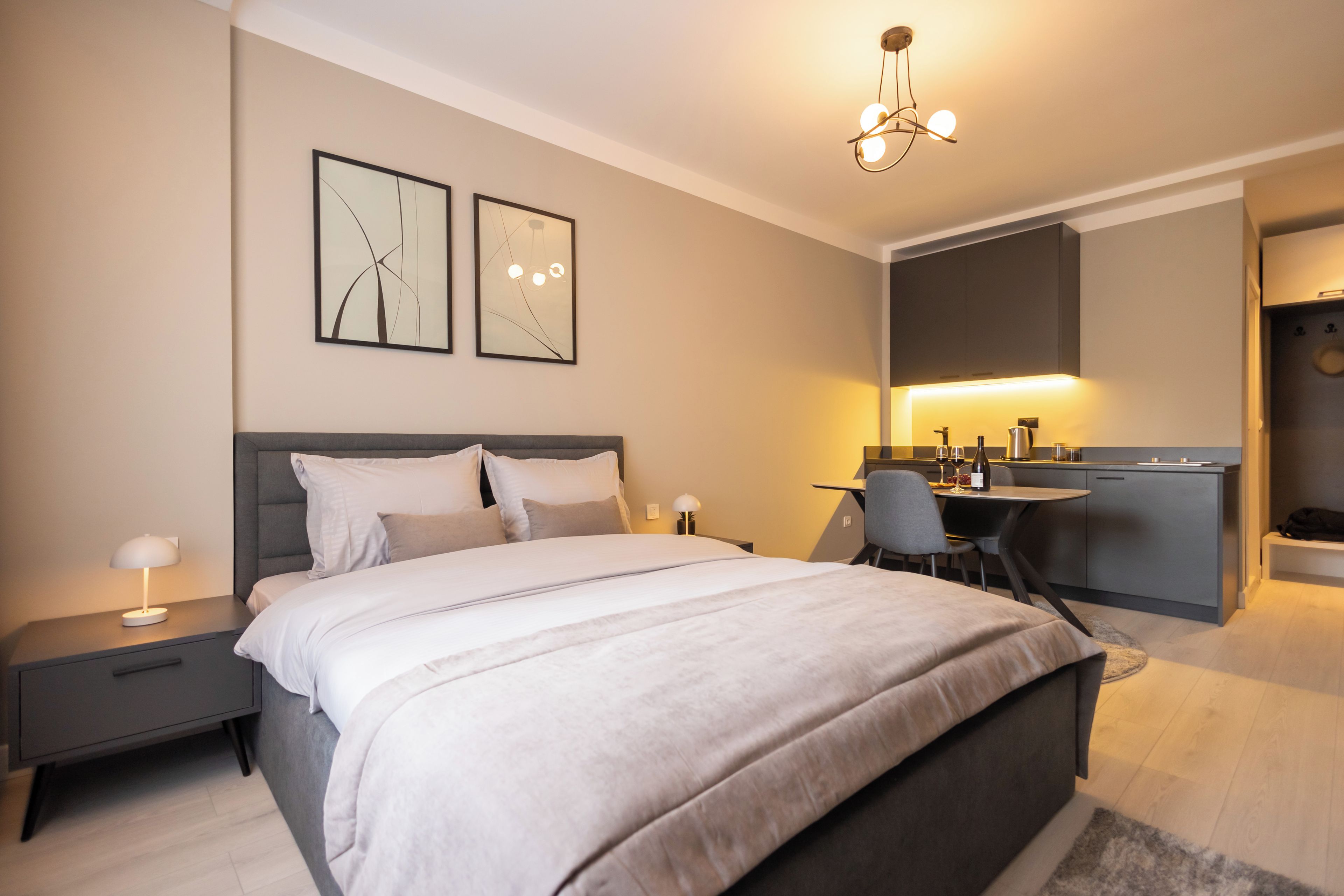 A modern bedroom with a large bed, bedside tables, a desk, and a kitchenette.