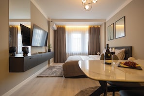Habitación de hotel moderna con una televisión de pantalla plana, una cama, una mesa de comedor con una botella de vino y uvas, y un ventanal con cortinas.